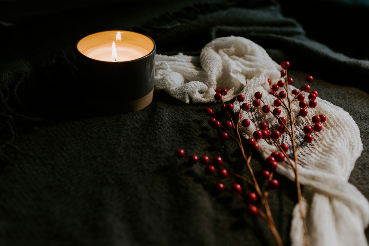 A lit candle in a dark ceramic holder beside a cream knit throw and sprigs of red winter berries.