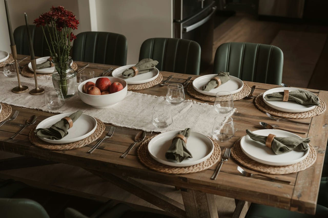 A wooden dining table set for eight with white plates, green linen napkins, a runner and a bowl of apples.