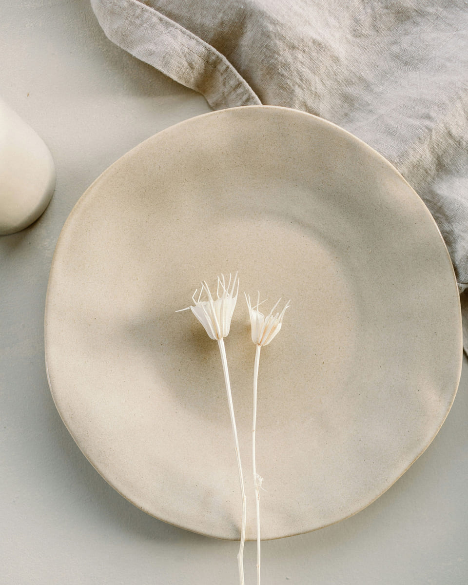 A hand-thrown cream stoneware plate with an organic edge, holding two dried white flowers, on linen.