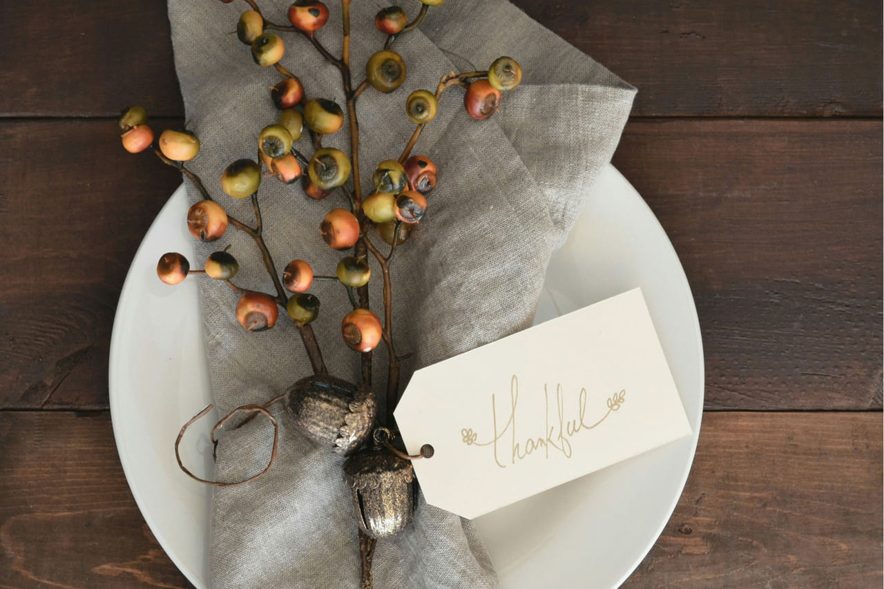 An overhead place setting: white plate, folded grey linen napkin, a sprig of autumn berries and a small card that reads Thankful.